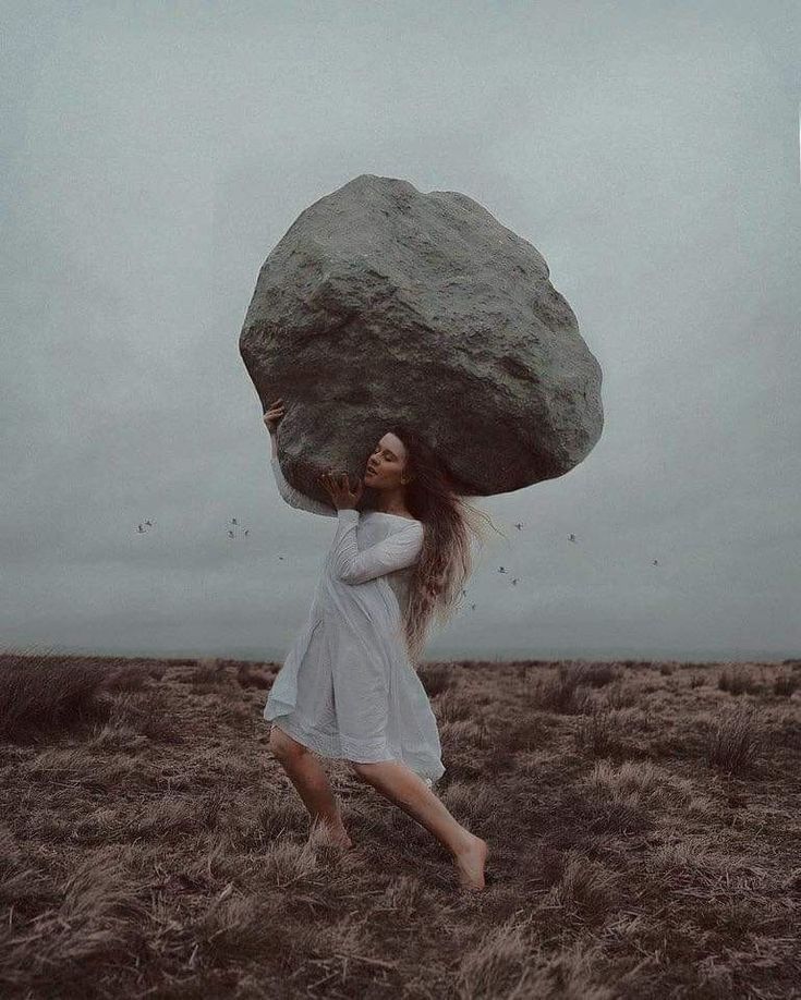 A figure carrying a boulder larger than themselves across an open field, sky overcast